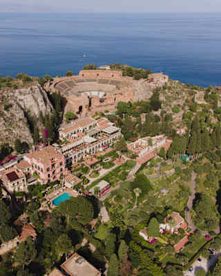 Aerial view over Grand Hotel Timeo with pool and exquisite gardens, to the ancient hilltop amphitheatre and azure sea beyond.