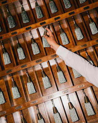 Angled image of a vintage wood key rack with numbered cubby holes, as a hand reaches for one of the keys with a tassel fob.