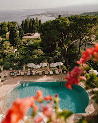 View over the pretty hotel pool, bordered by loungers and parasols, to gardens and the coast beyond, seen from a high window.