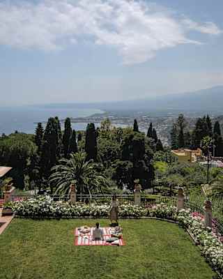 A couple enjoy a picnic in the lush gardens of the hotel, with views of the Ionian Sea, Naxos Bay and the slopes of Mt Etna