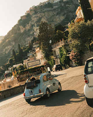 Angled image of a sky-blue Cinquecento with the top down, seen from behind as it travels a coast road bordered by cliffs.
