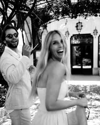 A newlywed woman with long blonde hair and her groom, both in white, laugh as they look back at the camera, in monochrome.