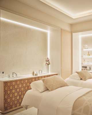 Two beds with beige and white bedding await guests in a room with backlit walls, one with basins and the other, product shelves.
