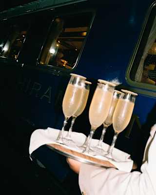 A person in a light-coloured suit holds a tray with four glasses of champagne in front of a dark railway carriage.