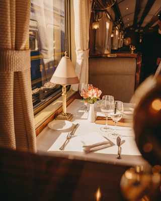 A dining table dressed with a white tablecloth, cutlery and glasses, adorned with a pink bloom in a vase, waits for guests.