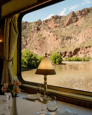 View from a table with glasses and a lamp through a window, where a slow, wide brown river cuts alongside red-stone cliffs.