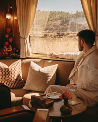 A man in pale clothing sits on a cushioned banquette with a cup of coffee gazing out of the sun-filled train window.