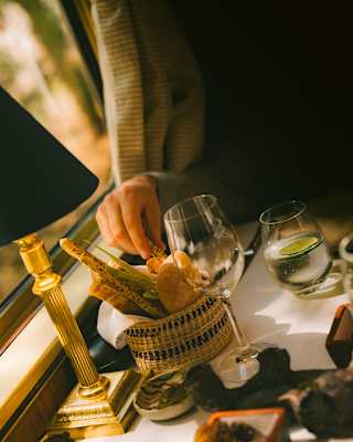 A diner's hand reaches for a seeded breadstick from a basket of rustic breads on a dining table with a wineglass and water.