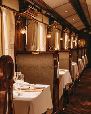 Elegant dining tables set with glasses and serviettes line the windowed aisle of a vintage railway carriage with warm lighting and plush seating, evoking a classic, luxurious travel atmosphere.