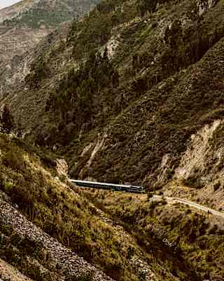The Hiram Bingham travels on a valley track between steep forested hillsides with rocky slopes, seen from above.