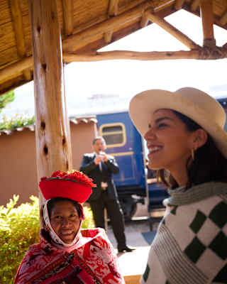 A woman in a sun hat and patterned poncho stands next to an Indigenous woman in traditional clothing and a red hat, with a railway carriage and a man in a suit in the background.