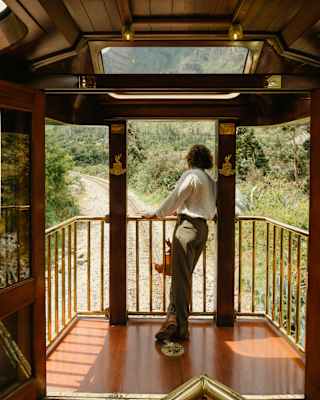 A man in trousers and white shirt leans on the railings, gazing at disappearing tracks, seen from inside the Observation Car.