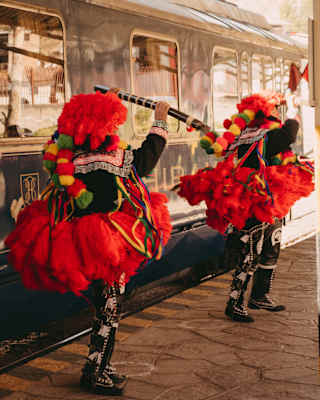 Two dancers in black trousers and flared red fluffy skirts, hats and pompoms, and holding flutes perform on a train platform.