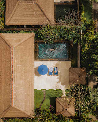 A guest floats in a sapphire pool of a Deluxe Villa, overlooked by a terrace with a parasol and loungers, seen from above.