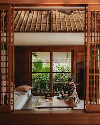 A woman kneels alongside a coffee table and prepares drinks on a tray, seen through beautiful open wood-slat windows.