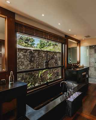 View of a villa bathroom, with floor, storage and frames in natural wood, with a long stone bath and grey-tiled shower area.