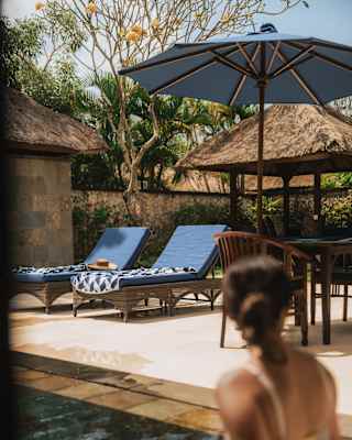 In soft-focus, a woman sits on the pool's edge, head turned to look at two blue sun loungers with towels, seen from behind.