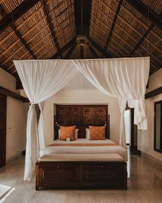 A high gabled ceiling with exposed beams rises above a four-posted bed with white drapes in the airy villa bedroom.