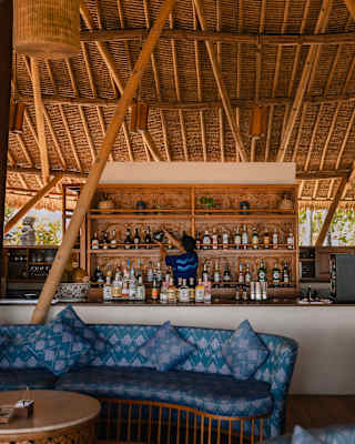 A thatched roof with giant supports towers above blue banquettes by the Puri Bar, where a barman is sorting the bottle shelf.
