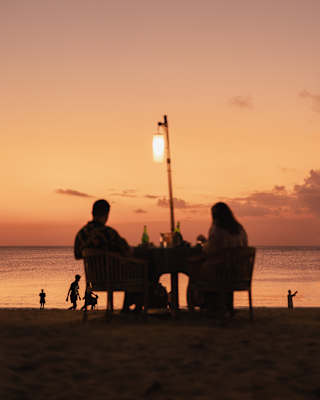 A silhouetted couple sits at a beachside table with a lantern, watching children play by the sea against a peach sunset sky.