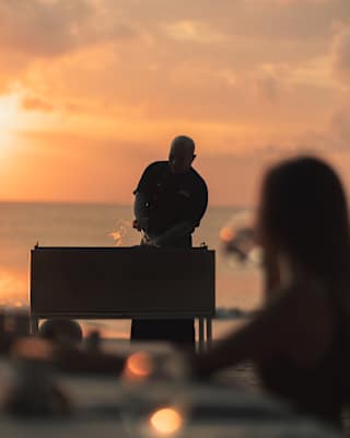 Silhouetted against the peachy sunset, a chef prepares food on a beach barbeque watched by a woman in soft-focus foreground.