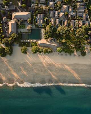 The foam edge of the blue ocean washes over the white beach outside the resort, dappled with early sun, seen from above.