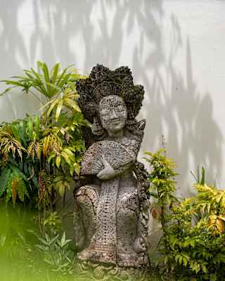 An ornate carved ancient stone statue of a smiling figure holding a fan sits in a quiet corner of the garden by a white wall.
