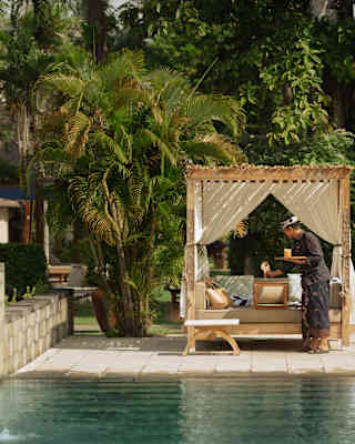 A staff member delivers drinks to a poolside cabana with cushions and drapes and a garden backdrop, seen across the pool.