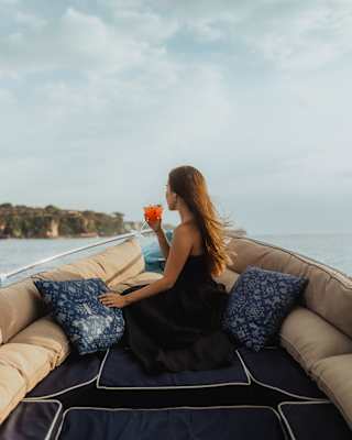 A long-haired woman in a black dress sits among cushions on the Dedari, holding an orange drink and gazing at the coast.
