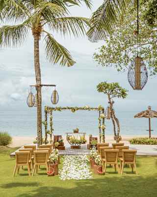 An aisle carpeted with white petals divides two small seating areas facing a flower wedding arch on lawn overlooking a beach.