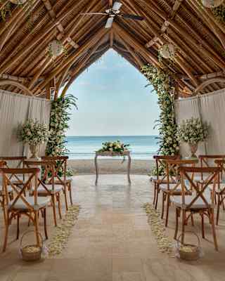 A petal-edged aisle divides chairs facing a registry table in an open-sided seaside venue with a pitched bamboo roof and drapes.
