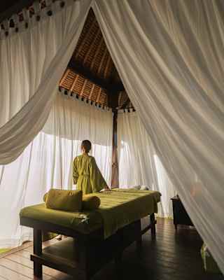 A woman in an olive-green gown stands by a treatment bed at the Beach Spa, seen from behind, through open white drapes.