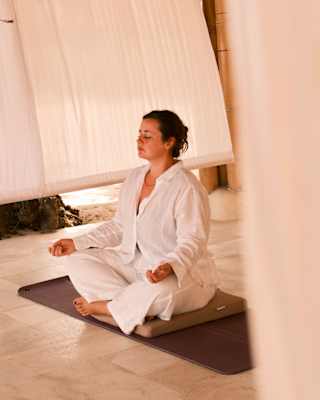 A woman in a loose-fitting white top and trousers sits on a black mat in lotus position with her eyes closed, in a cool room.