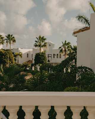 View of white Mediterranean-style villas rising from lush gardens, brushing the cloudy sky, seen from a terrace.