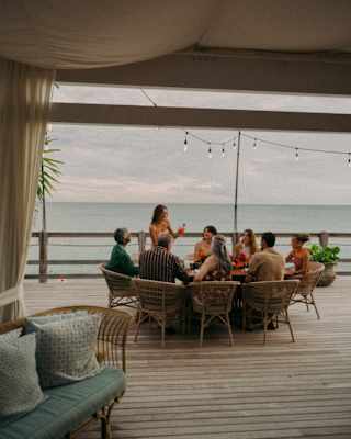 A woman stands and raises a toast watched by seven other guests seated at a round table on a deck overlooking the sea.