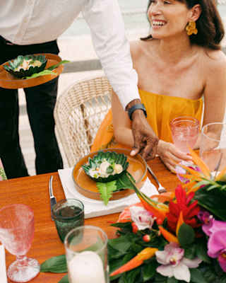 A waiter serves food in a delicate leaf-shaped ceramic green bowl to a dark-haired female guest in an orange top.