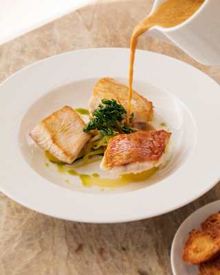 A jug pours a creamy sauce onto a plate of crisp-skinned white fish, with sautéed greens and herb oil, seen in close-up.