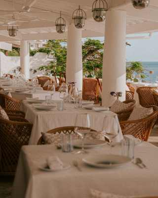 View across rows of laid tables with white cloths and wicker chairs in the shade of L'Oursin's covered sea-view terrace.