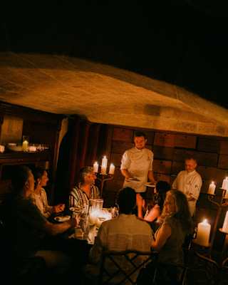 Angled image of two chefs bringing plates to a group of La Cave guests, dining by atmospheric candlelight in the cellar.