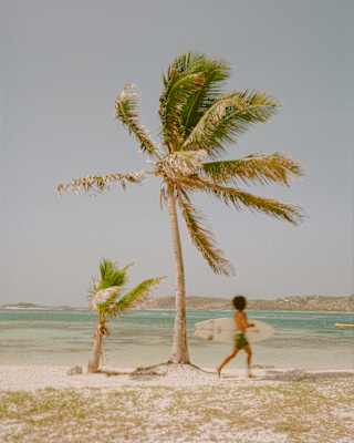A local man runs along sandy Galion Beach with a surfboard under his arm as wind whips the fronds of a nearby palm trees.