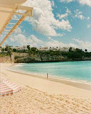 A figure stands at the edge of the sapphire sea, seen from beneath a canopy in a sun-drenched view of the beach and cliffs.