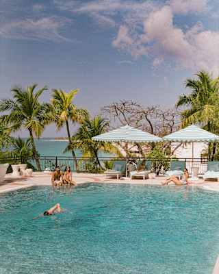 A man swims front-crawl across the pool, as three guests look on from the poolside, with a backdrop of palm trees and ocean.