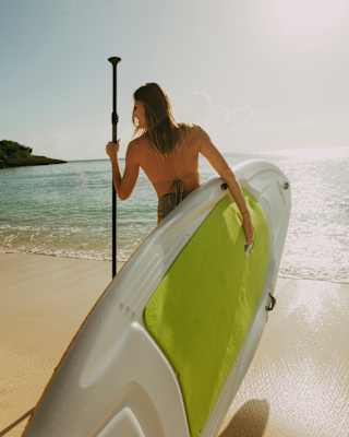 A woman holds a paddle in her left hand and a green and white board in her right, heading for the sea, seen from behind.