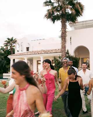 Six guests holding drinks and dressed up for an occasion laugh as they walk across gardens towards the camera, in close-up.