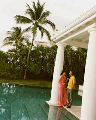 A man in a yellow shirt chats with a woman in a coral outfit by a pillar at the end of a terrace overlooking a still pool.