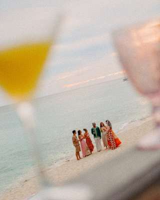 Six party-goers in bright clothing chat at the water's edge in an angled image, seen through cocktail glasses in soft-focus.