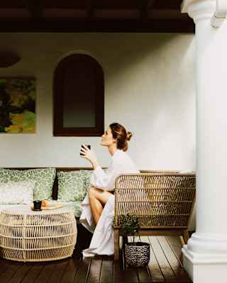 A woman in a white dressing gown sits on a wicker sofa outside, drinking from a mug. She is on a covered verandah with cushions, a small table, and a potted plant next to her.