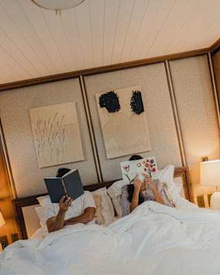 Two people are lying in bed under white duvets, each holding up a book to read. Their faces are hidden by the books, and the room has warm lighting with artwork on the wall above the bed.