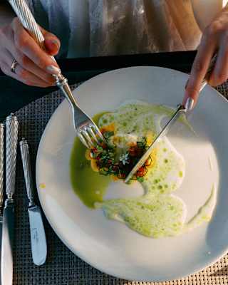 A person in a white top uses a fork and knife to cut into a gourmet dish with green sauce, served on a white plate on a woven placemat with silver cutlery.