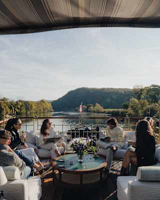 Five people sit and talk around a circular table with drinks on a boat deck, surrounded by water and trees, with a mountain visible in the background under a clear sky.